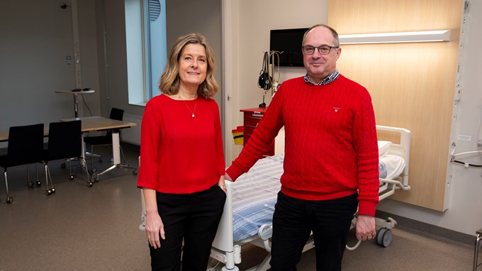 En kvinna och en man står vid en sjukhussäng, klädda i rött och svart.