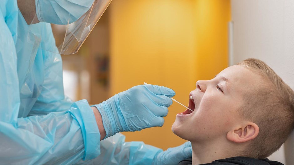 En ung pojke som testar sig för covid-19. Pojken har öppen mun och sjukvårdspersonal i skyddsutrustning provtar honom.