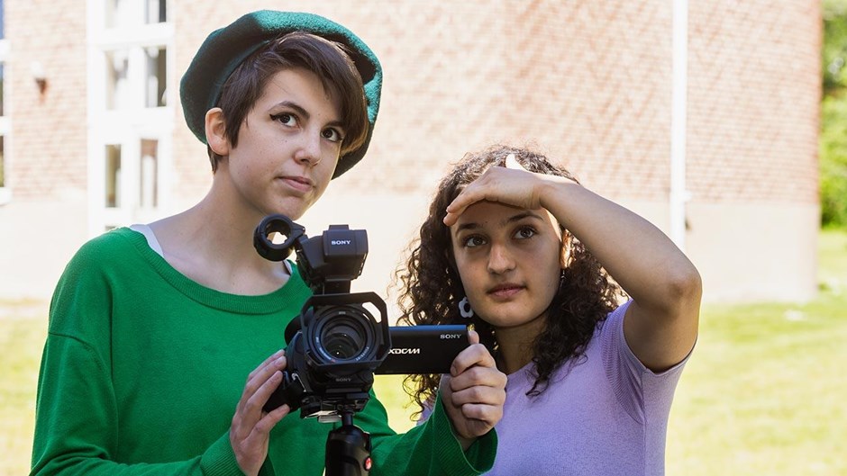 Två unga kvinnor står utomhus med en filmkamera. Den ena har grön tröja och en grönsvart mössa, den andrahar en ljuslila t-shirt. De tittar mot ett objekt vi inte ser, och den ena kvinnan håller upp handen till skydd mot solen.