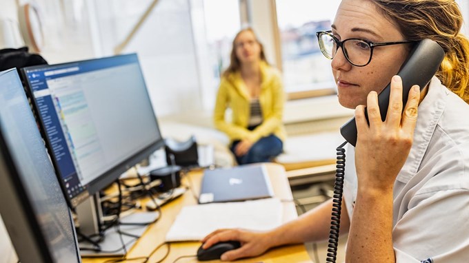 En kvinna i vårdkläder sitter med telefonlur i handen, medan en patient väntar i bakgrunden