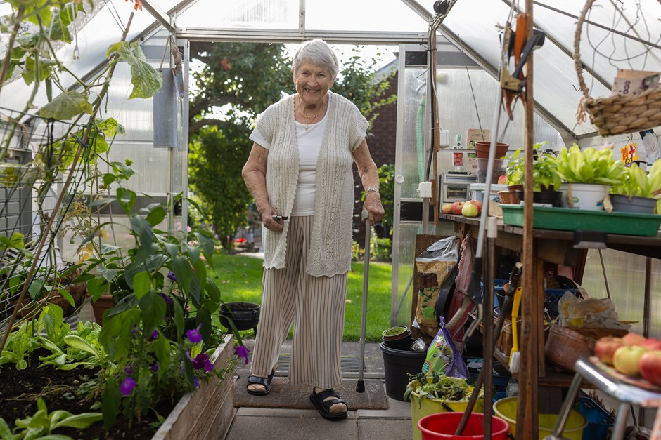En äldre dam med käpp i handen står i ett växthus med grönska runtomkring sig