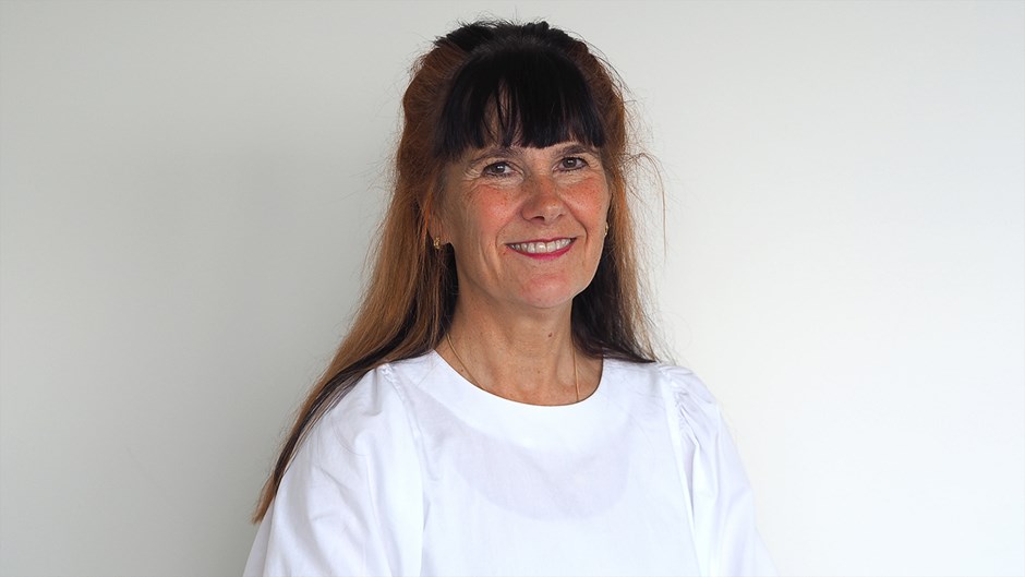 En kvinna med mörkt långt hår har en vit t-shirt på sig och står mot en vit vägg. Hon tittar rakt in i kameran och ler.
