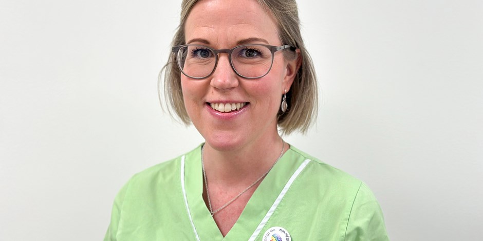 En kvinna i gröna klinikkläder och glasögon som tittar in i kameran och ler.