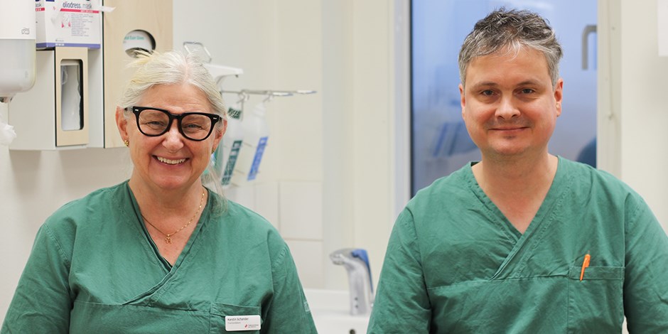 En man och en kvinna sitter i ett behandlingsrum. De har gröna klinikkläder på sig. De ler och tittar in i kameran.