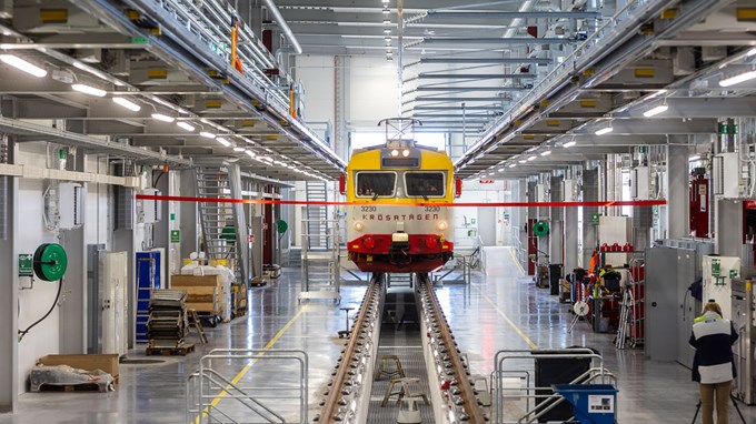 Ett gult tåg rullar in i en verkstadshall och har ett rött band hängande framför.