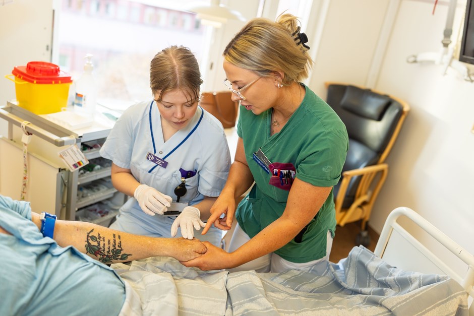 En sjuksköterska och en sjuksköterskestudent står vid en säng. Sjuksköterskan håller i en arm på en patient och pekar på den.
