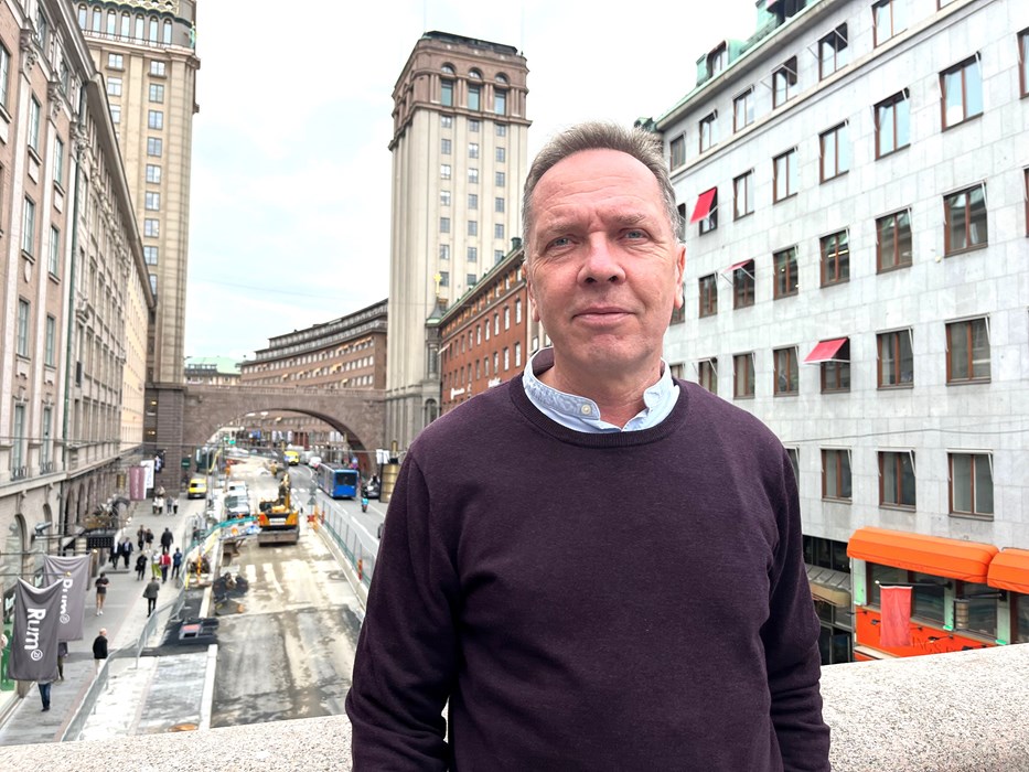 En man i blå tröja står på en gångbro över en stadsgata. Hus i bakgrunden.