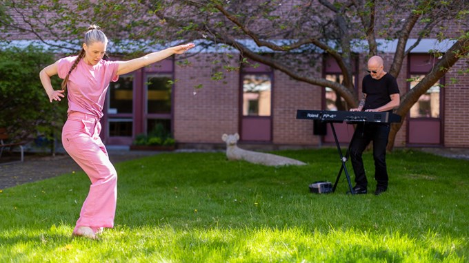 Kvinna i rosa kläder dansar medan en man i svart spelar på en synth i en trädgård.