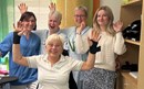 Fem kvinnor, fyra som står och en som sitter tittar in i kameran. Fyra av dem gestikulerar med armarna och de ser alla glada ut.