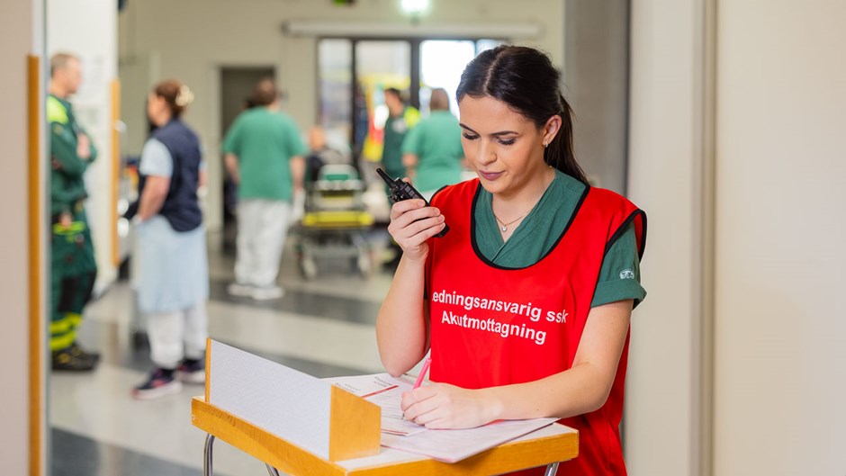 Kvinna med gröna sjukvårdskläder och en röd väst med texten ledningsansvarig ssk akutmottagning står vid en högt bord och håller upp en walkie talkie medan det är akutsjukvårdare i bakgrunden.