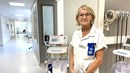 Kvinna står framför sjukhusutrustning. Hon är klädd i vita sjukhuskläder och bär glasögon.
