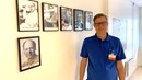 En man står vid en vägg och tittar in i kameran. Mannen är klädd i blå pikétröja och han bär glasögon. På väggen hänger fotografier av sex män som tidigare varit verksamhetschefer på infektionskliniken i Jönköping.
