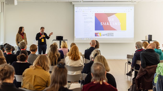 En person står i ett stort rum och talar inför ett antal åskådare.