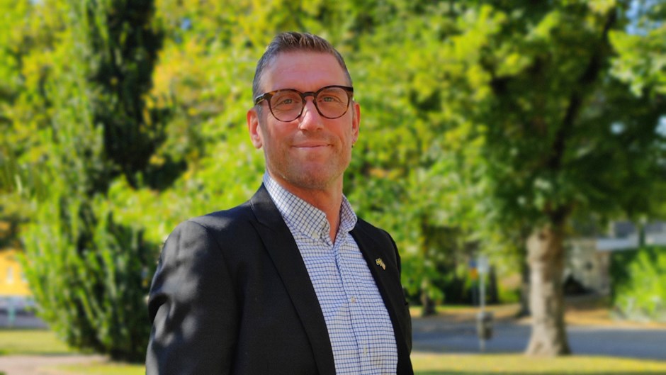 En man i skjorta och kavaj tittar in i kameran. Mannen bär glasögon. I bakgrunden flera grönskande träd.