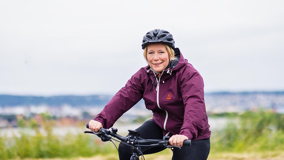 En kvinna i vinröd jacka och svart cykelhjälm cyklar med sjön Vättern i bakgrunden