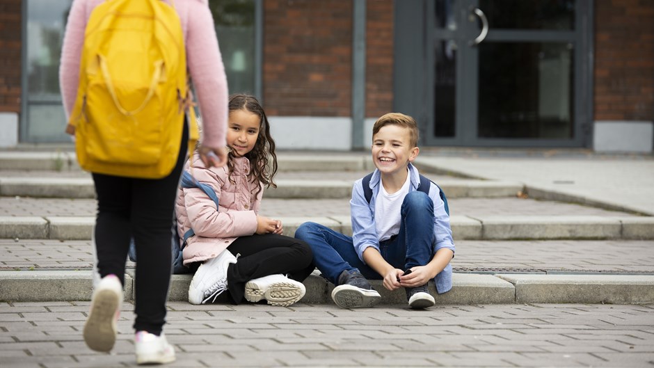 En pojke och en flicka sitter på ett betongsteg på en skolgård och tittar mot en skolkompis som kommer gående.