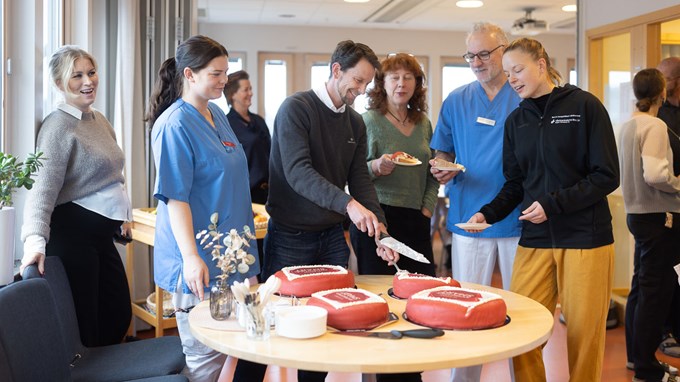 Fyra kvinnor och två män i ett fikarum på en vårdcentral står vid ett runt bord med fyra röda marsipantårtor och väntar på att få ta tårta.