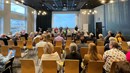 Många människor sitter på stolar i ett större konferensrum där deltagarna är vända fram mot scenen där det står tre män och pratar samt visar en presentation på storskärm.