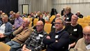 En kvinna står och pratar i en mikrofon mitt i ett rum med sittande publik, i ett konferensrum.