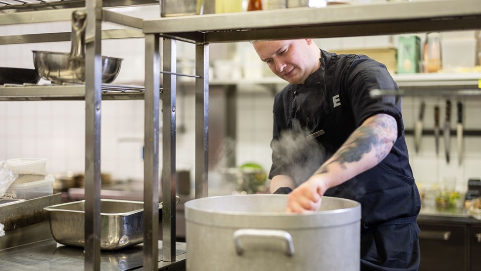 Ett restaurangkök med en ung man i svart kockskjorta som rör i en gryta.