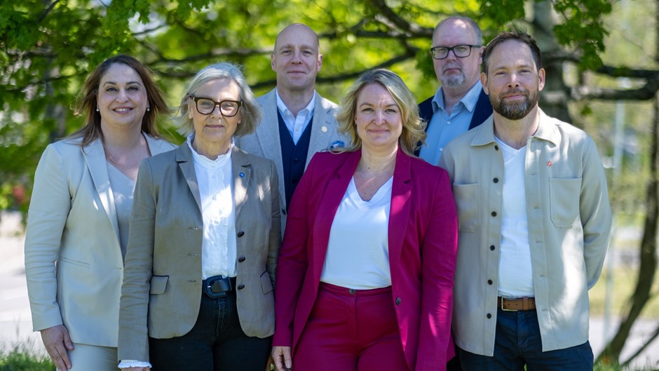 Tre kvinnor och tre män i somriga formella kläder står uppställda utomhus framför gröna buskar.