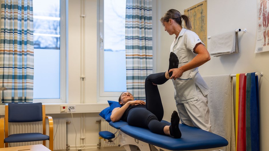 En kvinna i vårdkläder hjälper en annan kvinna på en bädd och testar hennes rörlighet i ena benet.