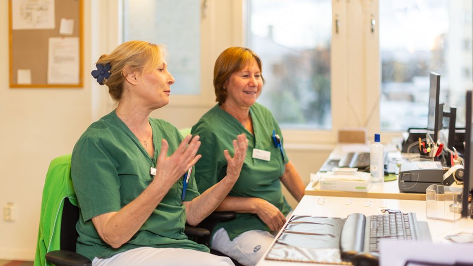 Två kvinnor i gröna sjukvårdskläder sitter på varsin skrivbordsstol vid ett skrivbord.