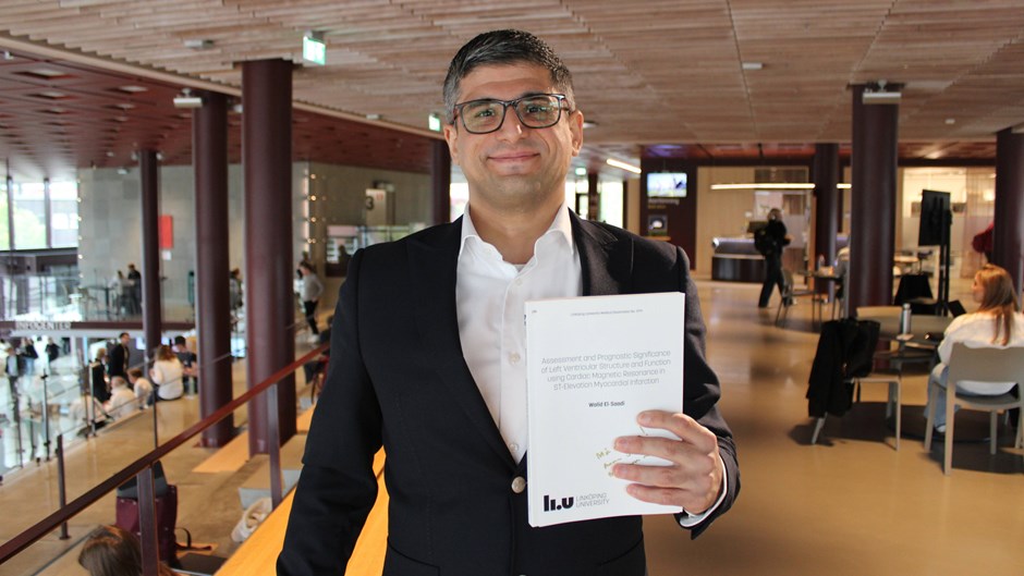 Man i glasögon och svart kavaj står i en rymlig lokal och håller upp sin doktorsavhandling.