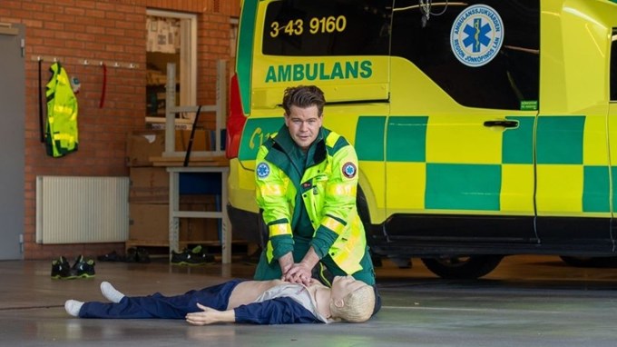En person utför hjärt- och lungräddning på en docka. I bakgrunden syns en ambulans.