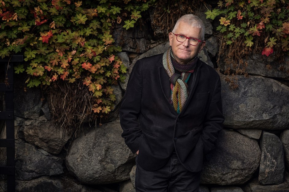 En man med kavaj, färgglad halsduk  och glasögon står framför en stenmur med höstfärgade löv.