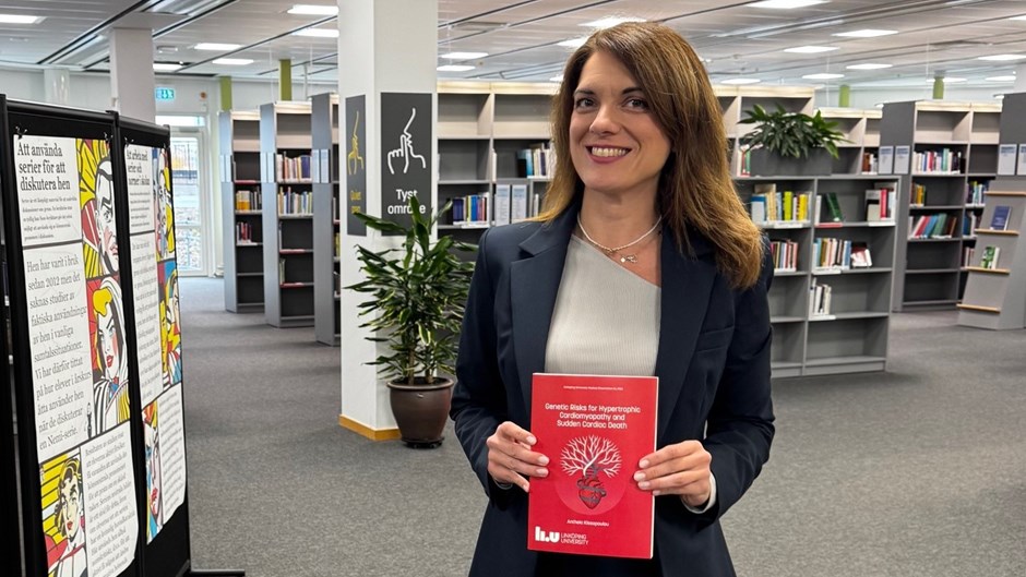 Kvinna i mörkt hår och mörk kavaj står och ler i ett bibliotek och håller upp en doktorsavhandling.