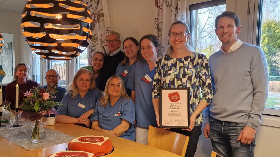 Människor, en del med sjukvårdskläder, står uppradade vid ett bord med flera tårtor och en kvinna håller i ett diplom.