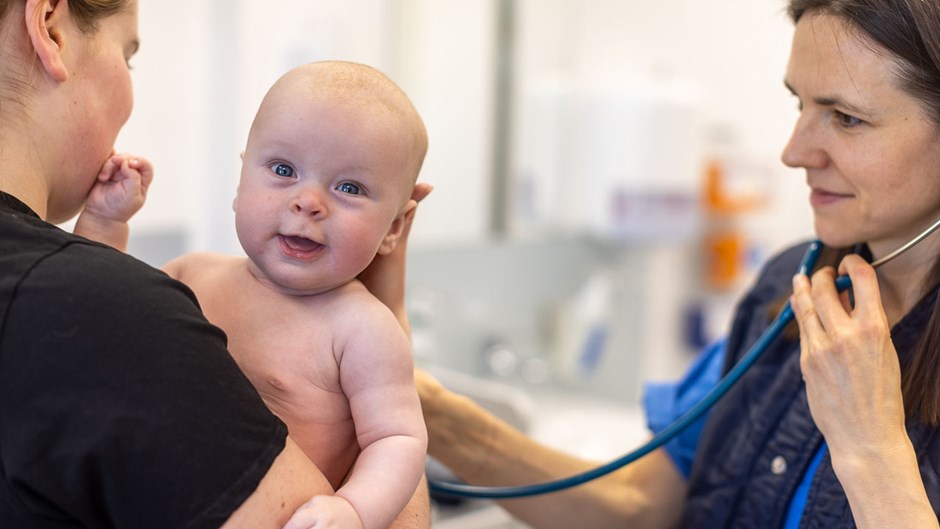 En läkare undersöker ett spädbarn, som barnets mamma håller i sin famn. Den kvinnliga läkaren har ett stetoskop runt halsen.