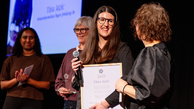 En mörkhårig kvinna med glasögon tar emot pris på scen av tre andra kvinnor.