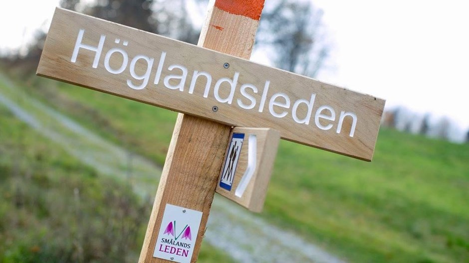 Träskylt med orange markering där det står Höglandsleden.