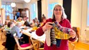 En kvinna i röd kofta håller upp stickade sockar i olika färger. I bakgrunden sitter flera kvinnor och stickar runt ett långt bord.