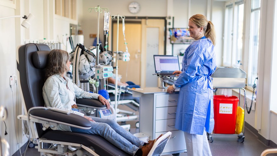 Två personer i vårdmiljö. En sitter i vad som ser ut som en patientstol och en står iförd vårdkläder. De båda pratar med varandra.