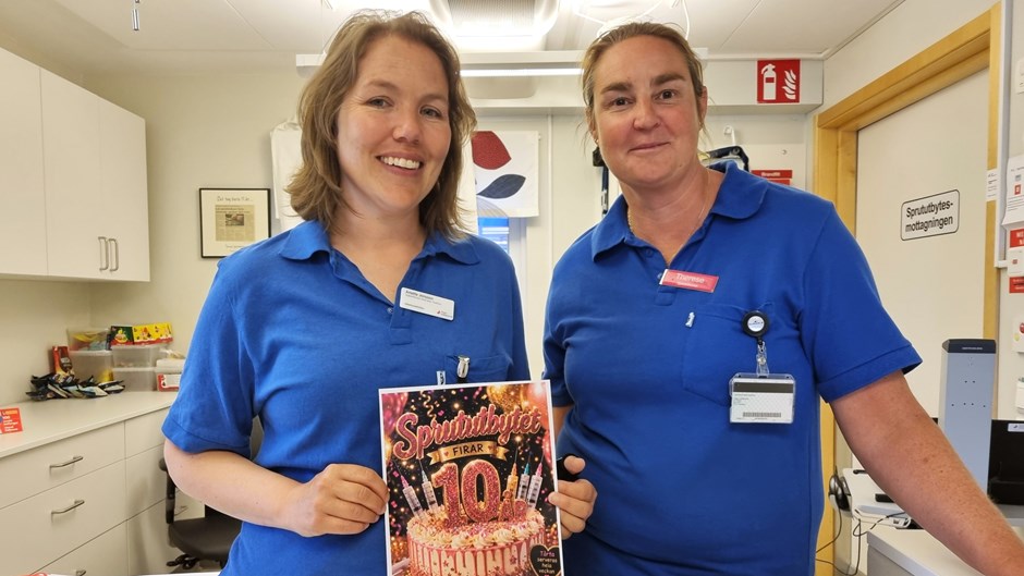 Två kvinnor i blåa vårdkläder håller upp en affisch om ett tioårsjubileum.