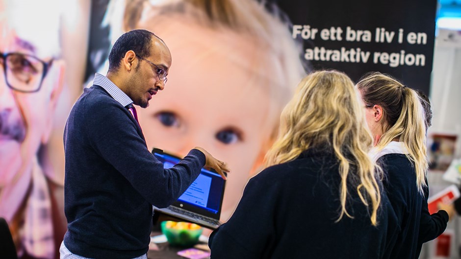 En man med laptop i händerna förklarar något för två personer med långt hår som står med ryggen mot kameran. I bakgrunden syns monterns väggar med bilder på en äldre man och ett litet barn.