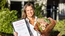 En leende kvinna håller upp ett inramat dokument och en blomsterbukett
