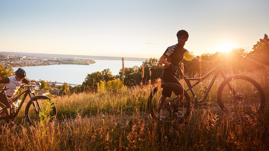 Två personer leder varsin cykel i högt gräs under en solnedgång.